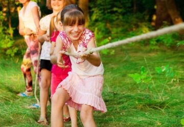 Group of happy children playing tug of war outside on grass. Kids pulling rope at park. Summer camp fun